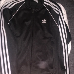 Svart Adidas track jacket med vita ränder - Svart Adidas track jacket med klassiska vita ränder längs ärmarna och Adidas logga på bröstet. Jackan har hel dragkedja framtill, ribbade muddar och är långärmad. Materialet är mjukt och glansigt, troligen polyester. Perfekt för en sportig och chill stil.