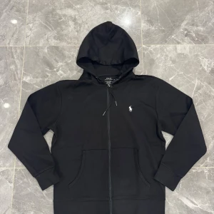 Svart hoodie från Polo Ralph Lauren - Svart hoodie från Polo Ralph Lauren med klassisk broderad logga på bröstet. Tröjan har huva med snörning, dragkedja framtill och två fickor. Tillverkad i mjuk bomull, perfekt för chill dagar. Snygg och enkel design som funkar till allt.