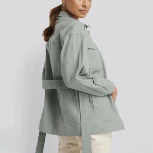 Ljusgrön tyg jacka med bälte - Snygg ljusgrön tyg jacka med knappar framtill, två bröstfickor med lock och ett långt bälte i midjan. Jackan har klassisk krage och raka, långa ärmar. 