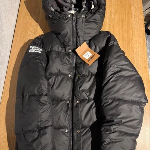 Svart pufferjacka från Burberry M - Svart pufferjacka från Burberry med avtagbara ärmar, huva och dragkedja, samt tryckknappar framtill. Klassisk Burberry-rutig insida i grått, vitt och rött. Patch med 'Burberry London England' på ärmen. Jackan har två sidofickor med dragkedja och quiltad design.