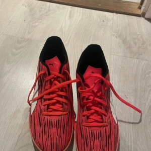 Röda Puma fotbollsskor med mönster - Säljer ett par röda Puma fotbollsskor med svart grafiskt mönster och vita loggor på sidan. Skorna har snörning och platt sula med grepp, perfekt för inomhusspel. Snygg design som sticker ut på planen.