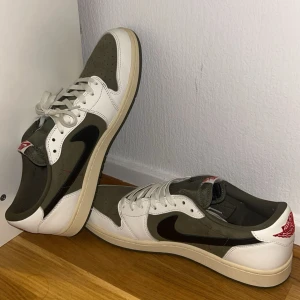 Nike Air Jordan 1 Low Travis Scott - Nike Air Jordan 1 Low Travis Scott med olivgröna och vita paneler, svart omvänd swoosh och röda broderade detaljer på hälen. Skorna har snörning, perforerad tå och en beige sula.