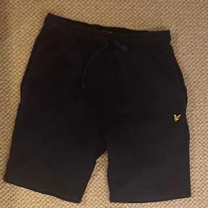 Svarta shorts från Lyle & Scott Junior - Svarta shorts från Lyle & Scott Junior med klassisk gul logga på ena benet. Mjukt material och bekväm passform med snörning i midjan. Perfekta för chill dagar eller träning. Snygg och enkel design som alltid funkar.