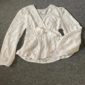 Vit omlottblus med knyt och spets - Söt vit omlottblus med knyt i midjan och långärmade puffärmar. Blusen har ett broderat blommönster i tyget och är tillverkad i ett tunt, lätt material. Perfekt för vår och sommar när du vill ha något luftigt och stilrent. Aldrig använd!