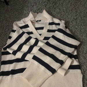 Randig stickad tröja från 157 - Snygg stickad tröja från 157 i storlek S. Den är vit med mörkblå breda ränder, har v-ringad hals och ribbade muddar vid krage, ärmslut och nederkant. Perfekt för dig som gillar klassisk stil med en modern twist.