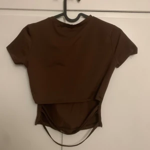 Brun croppad topp från MajorMade - Säljer en brun croppad topp från MajorMade med korta ärmar och rund hals. Toppen har en unik omlott-detalj nedtill med snörning baktill som ger en cool look. Materialet är lite träningsajtigt, mjukt och stretchigt, perfekt för dig som gillar trendiga och bekväma plagg.