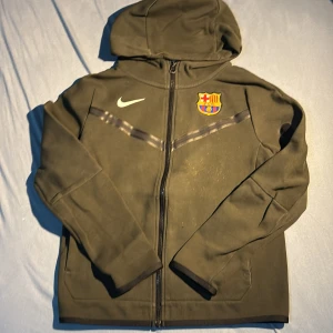Grön Nike FC Barcelona vindjacka - Snygg grön vindjacka från Nike med FC Barcelona-logga på bröstet och ryggen. Jackan har huva, hel dragkedja framtill och en extra ficka med dragkedja på ärmen. Perfekt för dig som gillar fotboll och vill ha en sportig look.