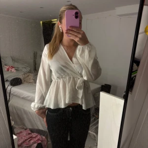 Vit omlottblus från Missy - Säljer en vit omlottblus från Missy med knyt i midjan och volang nedtill. Blusen har långärmade ärmar med lätt puff och v-ringad hals. Materialet är mjukt och har en lätt glansig finish som ger en fräsch look. Perfekt att styla med jeans eller kjol.