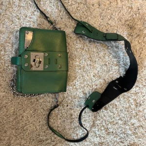 Grön axelväska med kedja och metall - Cool grön axelväska i skinn med bred axelrem och silverfärgade metalldetaljer. Väskan har ett unikt lås framtill och kedja som detalj. Insidan är svart och har plats för det viktigaste. Perfekt för dig som gillar edgy stil och vill sticka ut. Sytt ihop en trasig grejs se bild