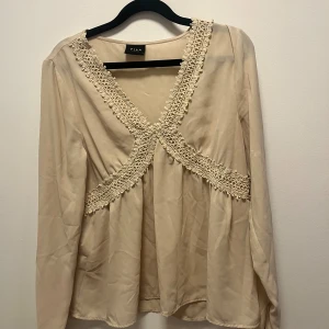 Beige blus med spets från Vila - Supersöt beige blus från Vila med v-ringning och dekorativa spetsdetaljer längs halsringningen och över bysten. Långärmad modell i mjukt, lätt material som ger ett flowy intryck. Perfekt för dig som gillar romantiska och feminina vibes.