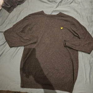 Mörkgrå stickad tröja Lyle & Scott - Snygg mörkgrå stickad tröja från Lyle & Scott med klassisk gul broderad fågel på bröstet. Tröjan har rund hals och långa ärmar, perfekt för dig som gillar stilrena och tidlösa plagg. Ribbstickade muddar vid ärmslut och nederkant ger extra komfort.