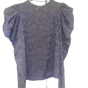 Svart blus med puffärm från Zara - Säljer en svart blus från Zara i storlek XS med subtilt blommönster och puffiga axlar. Blusen har en elegant, något loose passform och är tillverkad i ett lätt material som ger en lyxig känsla. Perfekt för dig som gillar statement-ärmar och diskreta detaljer.