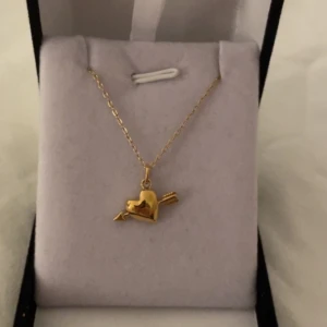 Guldhalsband med hjärta och pil litet - Snyggt halsband i guld med ett hängsmycke format som ett hjärta genomborrat av en pil. Kedjan är tunn och elegant, perfekt för att ge en romantisk touch till din stil. Hängsmycket har en blank finish och är riktigt gulligt. Rostfri