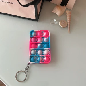 Säljer en multi-färgad pop it-nyckelring - Bilderna visar en färgglad pop it-nyckelring i rosa, blå och vit färg. Produkten är i gott skick utan synligt slitage eller defekter. Perfekt som stressleksak eller fidget för nyckelknippan. Lätt att ta med och använda när som helst.
