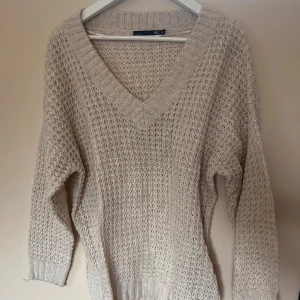 Beige stickad tröja från 157 - Mysig beige stickad tröja från 157 med v-ringning och grovstickat mönster. Tröjan har långa ärmar och breda ribbade muddar vid ärmslut och nederkant. Perfekt för lager-på-lager under kyliga dagar.
