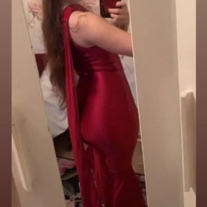 Röd asymmetrisk långklänning i satin - Säljer en röd, glansig långklänning i satin med asymmetrisk one-shoulder design och lång scarf-detalj (((FRÅN OHPOLLY))). Klänningen är tight och figurnära med volang nertill och en snygg, elegant siluett. Perfekt för dig som vill sticka ut med en unik look. Använd bara en! gång!.