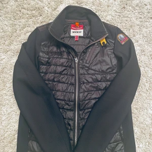 Svart quiltad jacka Parajumpers Y-L - Svart jacka från Parajumpers med quiltad front och rygg, samt släta ärmar. Jackan har dragkedja framtill, en gul detalj vid kragen och patch på axeln. Materialet är polyester och syntet, perfekt för kyliga dagar. Snygg och sportig look med modern passform. Barnstorlek motsvarar S 