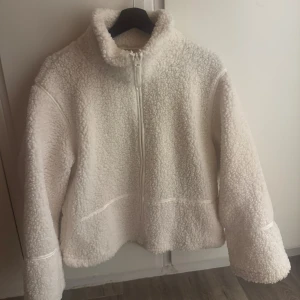 Vit teddyjacka med dragkedja - Mysig vit teddyjacka med hög krage och hel dragkedja framtill. Jackan har en fluffig och mjuk yta i fleece-material och är perfekt för kyliga dagar. Långärmad och med en avslappnad passform som ger en chill vibe.