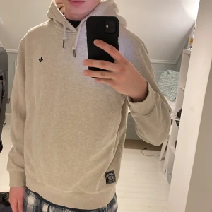 Morris hoodie - Snygg beige hoodie från Morris Stockholm i storlek M. Tröjan har huva med snörning, ribbade muddar och en liten svart broderad logga på bröstet. Materialet är mjuk bomull och insidan är fleecefodrad för extra komfort. Perfekt för chill dagar. Modell 178 cm. 