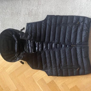 Mörkblå dunväst från Massimo Dutti - Mörkblå dunväst från Massimo Dutti med huva och horisontella sömmar. Västen har dragkedja framtill och två fickor med dragkedja. Perfekt för lager-på-lager och snygg till både jeans och hoodies.