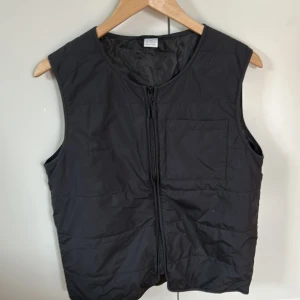 Svart dunväst från Lindex Kids - Svart dunväst från Lindex Kids med enkel design och rund hals. Västen har en praktisk dragkedja framtill och en bröstficka. Materialet är lätt och syntetiskt, perfekt för lager-på-lager under kyliga dagar. Passar dig som gillar stilren och funktionell look.