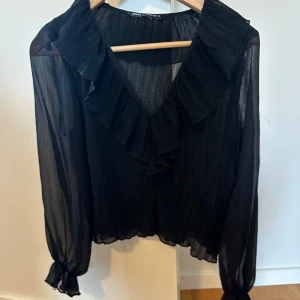 Svart blus med volang från Zara - Säljer en svart blus från Zara i tunn, transparent polyester. Blusen har långärmade ärmar med volangdetaljer vid mudden och en snygg v-ringad hals med bred volangkant. Perfekt för dig som gillar romantiska och trendiga detaljer.