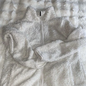 Vit teddyjacka med dragkedja - Mysig vit teddyjacka med hög krage och hel dragkedja framtill. Jackan har långärmade ärmar och är tillverkad i fluffigt teddy-material som ger en skön och chill vibe. Perfekt för kalla dagar när du vill vara både bekväm och snygg.