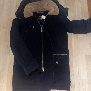 Snoot -           Stilren vinterjacka från Snoot i mörkblått med beige teddykrage, dragkedja och knappar framtill. Jackan har flera fickor, detaljer i metall och fodrad insida. Perfekt för kalla dagar med sin långa passform och varma material. Perfekt till vintern och sen höst. Passform: slimfit