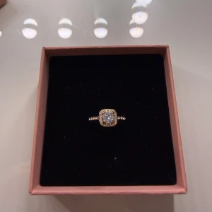 Guldfärgad ring med glittrande stenar - Snygg guldfärgad ring med fyrkantig infattning och en stor gnistrande sten i mitten, omgiven av mindre stenar runt om. Ringen har även små stenar längs bandet för extra bling. Perfekt för dig som gillar att sticka ut med dina accessoarer.