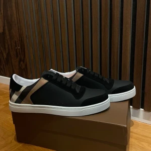 Burberry rutig sneakers svart/vit - Snygga Burberry sneakers med klassiskt rutigt mönster i beige, svart och vitt på sidan. Ovandelen är i svart skinn och mocka, med vita platta sulor och svarta snören. Perfekt för dig som gillar stilrena och lyxiga detaljer.