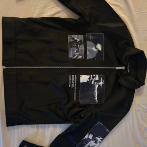 Svart vindjacka från Manière De Voir - Svart vindjacka med stora svartvita fotoprints på både fram- och baksida samt ena ärmen. Jackan har en rak passform, hel dragkedja framtill och coola detaljer som ger streetkänsla. Materialet är syntet och jackan har ståkrage. Perfekt för dig som gillar sportiga och unika plagg.