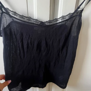 Svart linnetopp med spets från Massimo Dutti - Svart linnetopp från Massimo Dutti med tunna axelband och transparent spetskant med blommönster längs halsringningen. Toppens material är mjukt och lätt, perfekt för varma dagar. Snygg att styla med jeans eller kjol för en chill look.