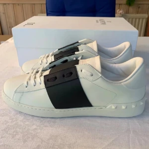 Valentino Garavani Open sneakers vit/svart - Snygga Valentino Garavani Open sneakers i vitt skinn med bred svart läderdetalj över mitten. Klassisk rund tå, vita snören och ikoniska nitar på hälen. Perfekta för dig som vill ha en stilren och lyxig look med en modern twist.