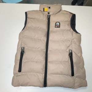 Beige dunväst från Parajumpers - Snygg beige dunväst från Parajumpers med svart dragkedja och coolt PJS-märke på bröstet. Västen har två praktiska fickor med dragkedja och en hög krage med gul detalj. Insidan är fodrad i svart och har flera etiketter med info om märket.
