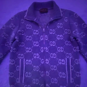 Gucci teddyjacka med GG-mönster - Säljer en lyxig teddyjacka från Gucci med ikoniskt GG-mönster över hela jackan. Jackan är blå med vita detaljer, har hög krage, dragkedja framtill och ribbade muddar vid kragen och fickorna. Perfekt statement-plagg för dig som gillar exklusiv streetstyle.