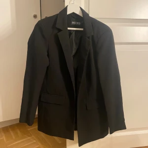 Svart kavaj från Pieces - Stilren svart kavaj från Pieces med klassisk krage och två fickor framtill. Rak passform och helsvart design, perfekt för en snygg och stilren outfit. Nypris 599kr.