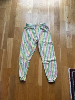 Randiga mjukisbyxor från Mini Rodini - Säljer ett par mjukisbyxor från Mini Rodini med färgglada ränder i pastellrosa, ljusgrön, gul och vit. Byxorna har resår i midjan och är gjorda i mjuk bomull. Perfekta för chill dagar och har broderad logga på sidan.