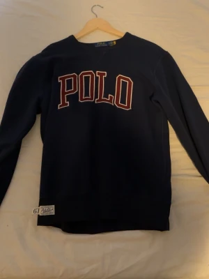 Mörkblå sweatshirt från Polo Ralph Lauren - Snygg mörkblå sweatshirt från Polo Ralph Lauren med stor vit och röd POLO-text på bröstet. Tröjan har långa ärmar, rund hals och en cool detalj med etikett och siffran 67 vid nederkanten. Perfekt för chill dagar och streetwear-stil.