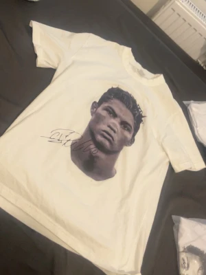 Vit t-shirt med porträtttryck - Säljer en vit t-shirt med coolt porträtttryck framtill och en tryckt autograf under motivet. T-shirten har klassisk passform, rund hals och är kortärmad. Materialet är mjuk bomull som känns skönt mot huden. Perfekt för dig som gillar unika och grafiska plagg.