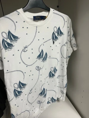 Vit t-shirt med segelbåtar från Polo Ralph Lauren - Vit t-shirt från Polo Ralph Lauren med marinblå segelbåtar, ankare och rep som mönster. T-shirten har korta ärmar och är gjord i mjuk bomull. Perfekt för dig som gillar nautisk stil och vill sticka ut med coolt print.