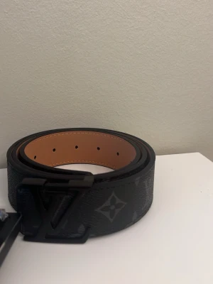 Svart L&V skinnbälte - Säljer ett svart L o v bälte i skinn med klassiskt monogrammönster och svart L & V spänne. Insidan är ljusbrun och bältet är 100 cm långt. Perfekt accessoar för att lyfta din outfit med en lyxig touch.