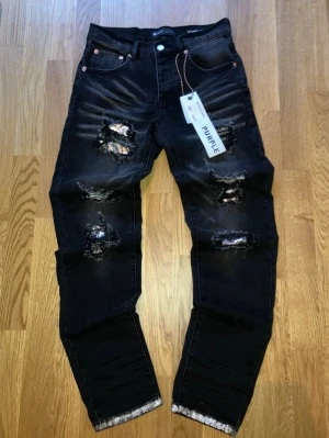 Svarta Purple Brand Jeans - Säljer ett par svarta jeans från Purple Brand i storlek 28 (165-175cm). Jeansen har slitna detaljer med glittriga inslag framtill och ett rutigt tyg på bakfickan. De har en tapered passform och är tillverkade i jeansmaterial med snygga dragkedjor och knappar.