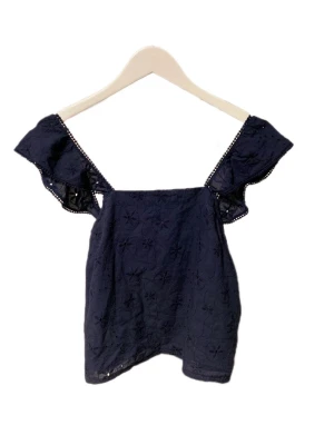 Mörkblå blus med volang från Pieces - Säljer en mörkblå blus från Pieces i storlek S. Blusen har breda axelband med volangdetaljer och broderat blommönster över hela tyget. Modellen är ärmlös och har en luftig passform, perfekt för varmare dagar.