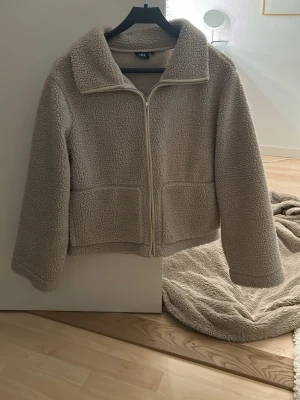 Beige teddyjacka från 157, storlek S - Mysig beige teddyjacka från 157 i storlek S. Jackan har en klassisk krage, två stora fickor framtill och stängs med dragkedja. Materialet är fluffigt och fleece-liknande, perfekt för kyliga dagar. Passformen är normal och jackan har långa ärmar.