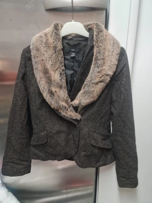 Brun kavaj med fuskpäls krage från H&M - Säljer en brun kavaj från H&M med bred, mjuk fuskpäls i beige runt kragen. Västen har ett diskret rutmönster och två fickor framtill. Materialet är ullblandning och polyester, vilket ger en lyxig känsla. Perfekt för dig som vill ha en cool och trendig look.