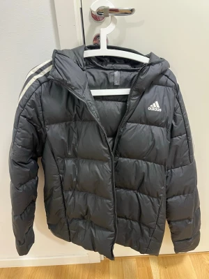 Svart pufferjacka från Adidas - Säljer en svart pufferjacka från Adidas med huva och dragkedja. Jackan har klassiska vita Adidas-ränder på axlarna och logga på bröstet. Perfekt för kalla dagar och har en schysst, sportig look. Materialet är polyester och jackan är långärmad.