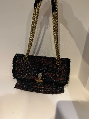 Svart axelväska från Kurt Geiger - Säljer en snygg svart axelväska från Kurt Geiger med grovvävt tyg i mörka toner och inslag av rött och blått och glitter.Väskan är använd och har tappat ett öga på örnen och har tyvärr ett hål i ena innefacket men fortfarande väldigt fin.