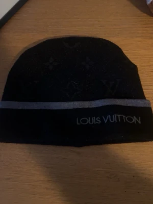 Svart mössa  - Svart mössa med diskret monogrammönster och en grå rand runt om. Märkets logga är broderad framtill. Mössan har en klassisk form och är tillverkad i ett mjukt material som känns lyxigt och bekvämt.