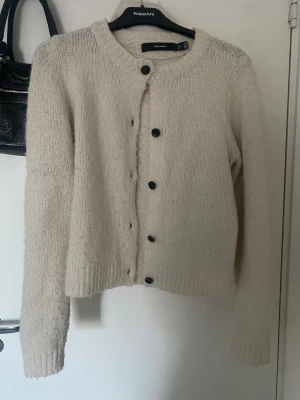 Beige stickad kofta från Vero Moda - Mysig beige stickad kofta från Vero Moda med rund hals och svarta knappar framtill. Koftan har långärmade och ribbade muddar vid ärmslut och nederkant. Perfekt för lager-på-lager under kyliga dagar.