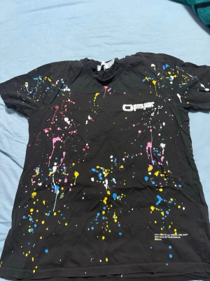 Svart Off-White t-shirt med färgstänk - Säljer en svart t-shirt från Off-White med coola färgstänk i blått, gult, rosa och vitt. Framsidan har Off-White-logga och baksidan har den ikoniska pil-designen. T-shirten är kortärmad och gjord i mjuk bomull, perfekt för dig som gillar streetwear och vill sticka ut.